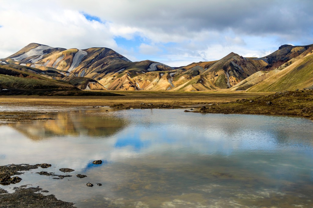 The rainbow rock of Landmannalaugar is reflected in a still lake under a cloudy sky in Iceland
